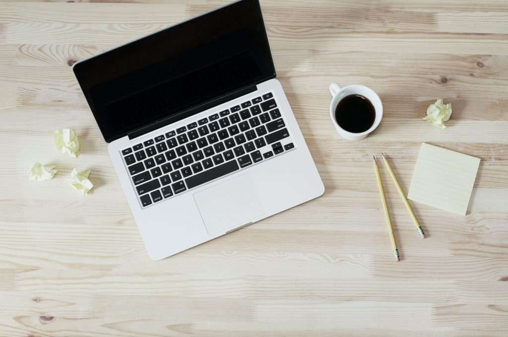 A laptop on a wooden table with a cup of coffee and a notepad.